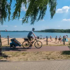 Rowerzysta z przyczepką jedzie ścieżką przy plaży nad Pogorią. Ludzie odpoczywają na piasku.