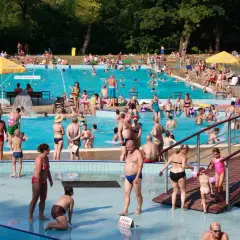 Tłum ludzi korzysta z basenu w upalny dzień. Wszyscy ubrani są w stroje kąpielowe, niektórzy w koszulki na basen, by chronić się przed słońcem.
