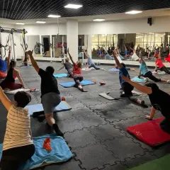 Grupa osób ćwiczy jogę na macie w siłowni Tychy Stadion. Lustra odbijają salę pełną ćwiczących.