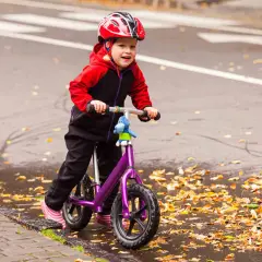 Mała dziewczynka w kasku uczy się jeździć na fioletowym rowerku biegowym. Jesienne liście i kałuże wokół.