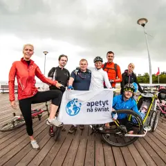 Grupa rowerzystów z banerem "apetyt na świat" pozuje do zdjęcia na molo, gotowi na wyprawę rowerem na Hel.