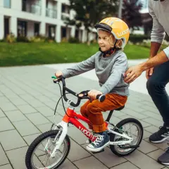 Chłopiec w kasku uczy się jeździć na czerwonym rowerze. Tata pomaga, pokazując, jak dobrać rower dla dziecka.