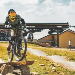 Rowerzysta w kasku zjeżdża po drewnianej kładce, przypominającej ścieżkę w kamieniołomie. W tle kolejka linowa.