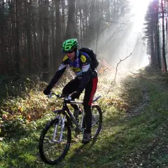 Rowerzysta w kasku jedzie leśną ścieżką, która jest częścią szlaku stu jezior. Mgła i promienie słońca tworzą magiczną atmosferę.