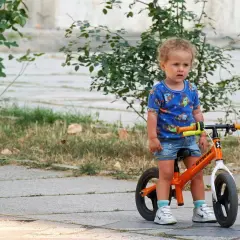 Małe dziecko w niebieskiej koszulce z pieskami stoi obok swojego pomarańczowego rowerka dla niemowlaka.