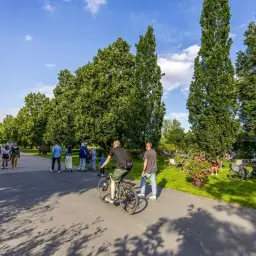 Ludzie korzystają z pięknej pogody na ścieżkach rowerowych w Poznaniu. W parku widać spacerowiczów, rowerzystów i osoby odpoczywające na trawie.