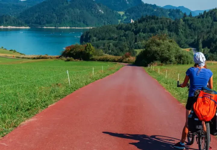 Kobieta na rowerze z bagażem na czerwonej ścieżce rowerowej. Piękna trasa Velo Dunajec z widokiem na jezioro i góry.