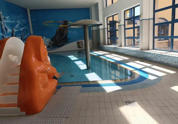 Wnętrze basenu z pomarańczową zjeżdżalnią, muralem z delfinami i dużymi oknami.