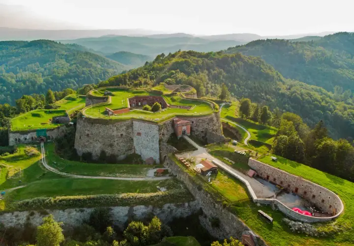 Forteca na wzgórzu, otoczona zielenią, z widokiem na góry. Srebrna góra trasa zaprasza do odkrywania historii.
