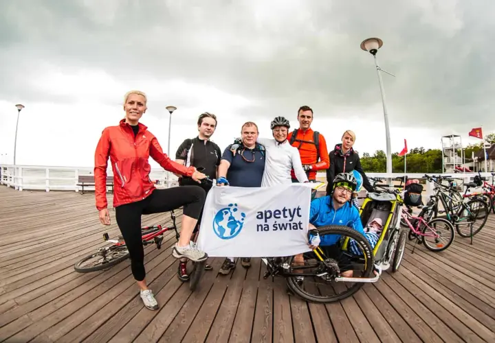 Grupa rowerzystów z banerem "apetyt na świat" pozuje do zdjęcia na molo, gotowi na wyprawę rowerem na Hel.