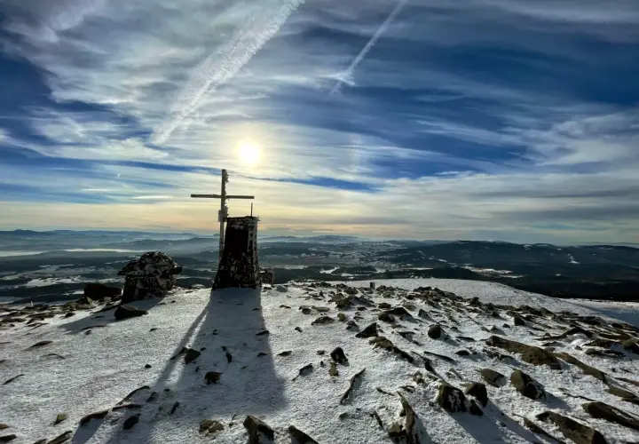 Szczyt Babiej Góry zimą. Krzyż i kamienna konstrukcja na ośnieżonym szczycie. W tle rozległa panorama gór i niebo z chmurami. Szlaki Babiej Góry w śniegu.