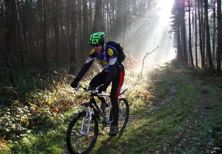 Rowerzysta w kasku jedzie leśną ścieżką, która jest częścią szlaku stu jezior. Mgła i promienie słońca tworzą magiczną atmosferę.
