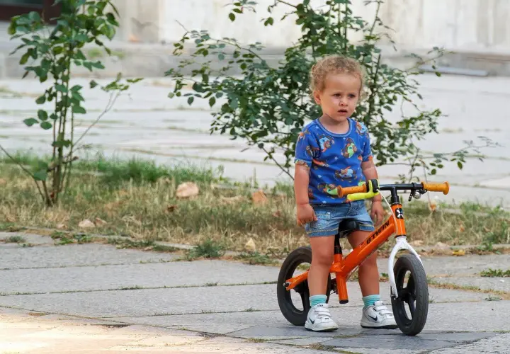 Małe dziecko w niebieskiej koszulce z pieskami stoi obok swojego pomarańczowego rowerka dla niemowlaka.