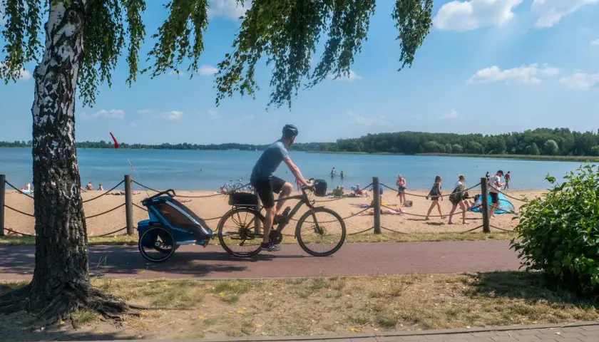 Rowerzysta z przyczepką jedzie ścieżką przy plaży nad Pogorią. Ludzie odpoczywają na piasku.