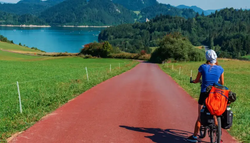 Kobieta na rowerze z bagażem na czerwonej ścieżce rowerowej. Piękna trasa Velo Dunajec z widokiem na jezioro i góry.
