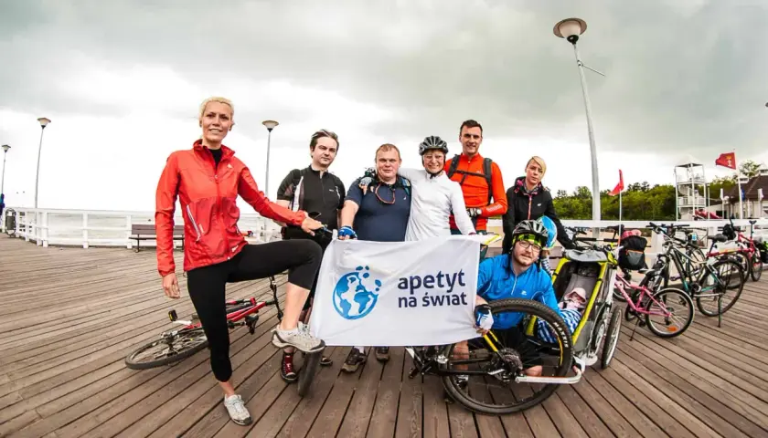 Grupa rowerzystów z banerem "apetyt na świat" pozuje do zdjęcia na molo, gotowi na wyprawę rowerem na Hel.