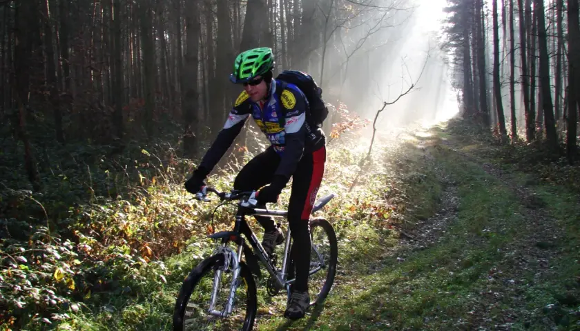 Rowerzysta w kasku jedzie leśną ścieżką, która jest częścią szlaku stu jezior. Mgła i promienie słońca tworzą magiczną atmosferę.