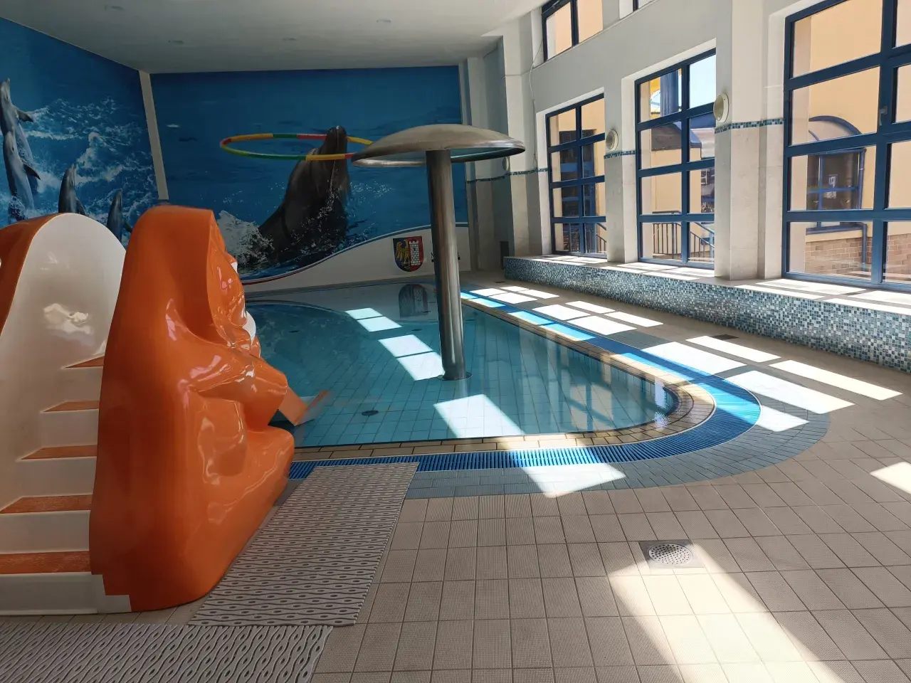 Wnętrze basenu z pomarańczową zjeżdżalnią, muralem z delfinami i dużymi oknami.