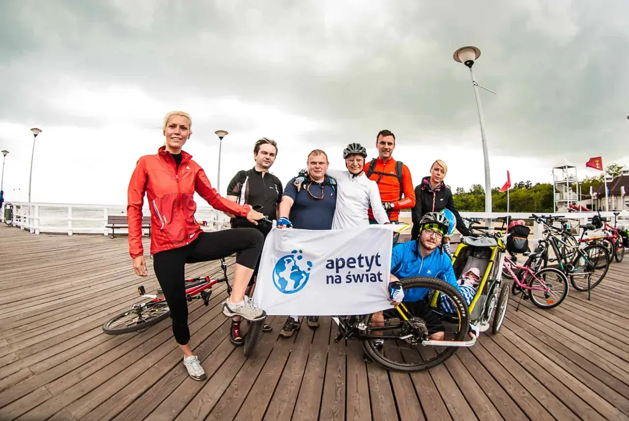 Grupa rowerzystów z banerem "apetyt na świat" pozuje do zdjęcia na molo, gotowi na wyprawę rowerem na Hel.