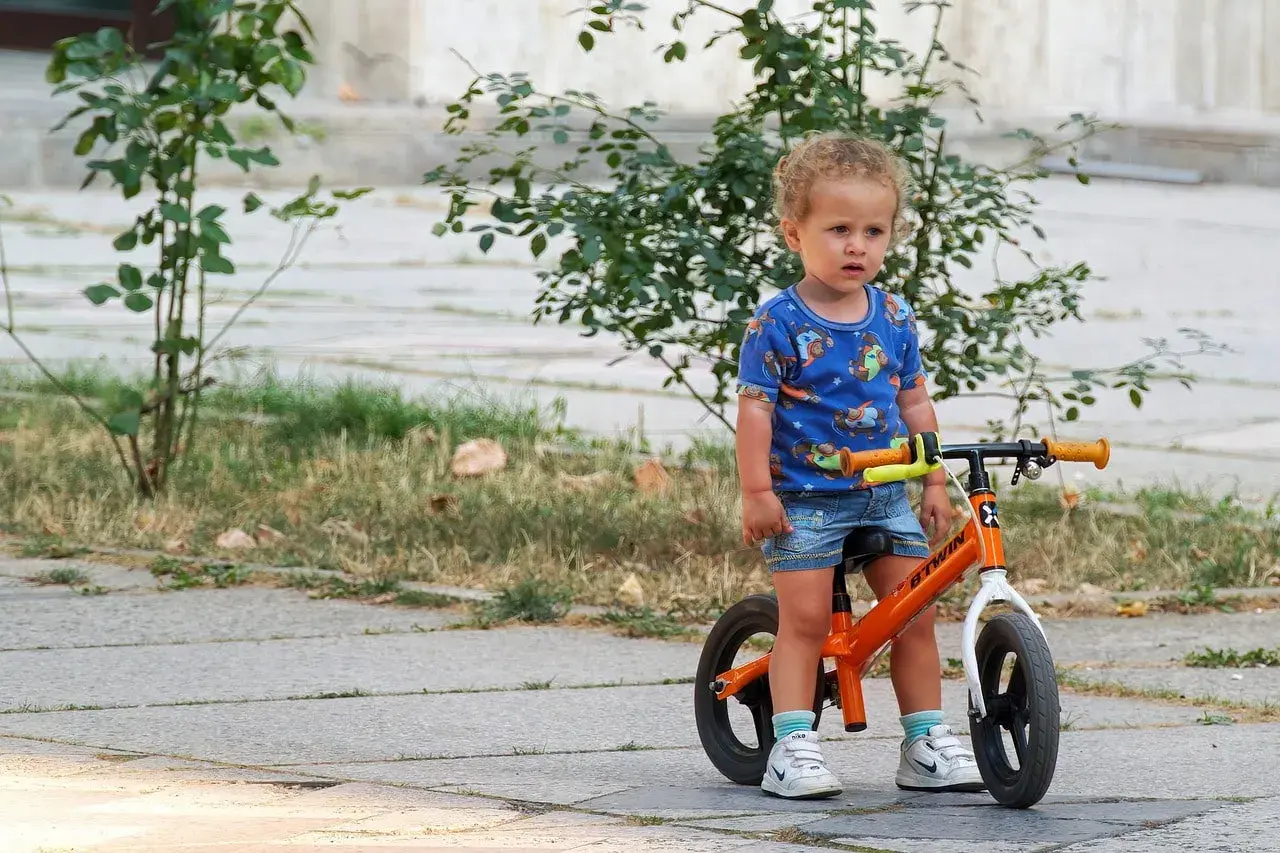 Małe dziecko w niebieskiej koszulce z pieskami stoi obok swojego pomarańczowego rowerka dla niemowlaka.