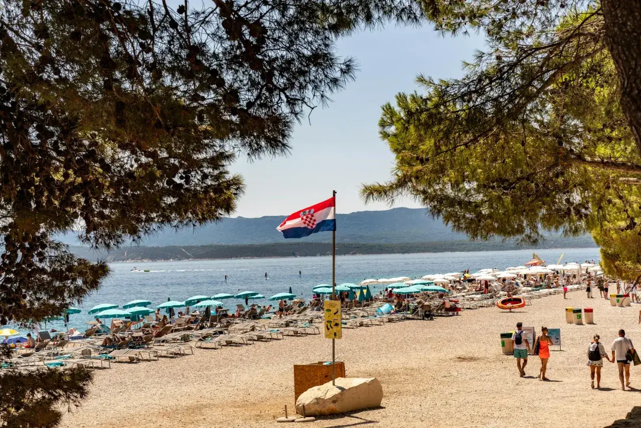Słoneczna plaża w Chorwacji, z chorwacką flagą powiewającą na wietrze. Ludzie odpoczywają na leżakach pod parasolami. Idealne miejsce na wakacje, gdzie można zapomnieć o wszystkim i cieszyć się słońcem.