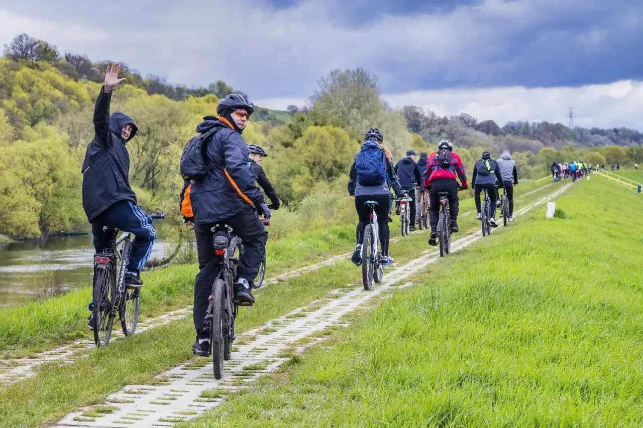 Grupa rowerzystów przemierza malowniczą wiślaną trasę rowerową. Wiosenna zieleń otacza ścieżkę, a chmury zapowiadają deszcz.