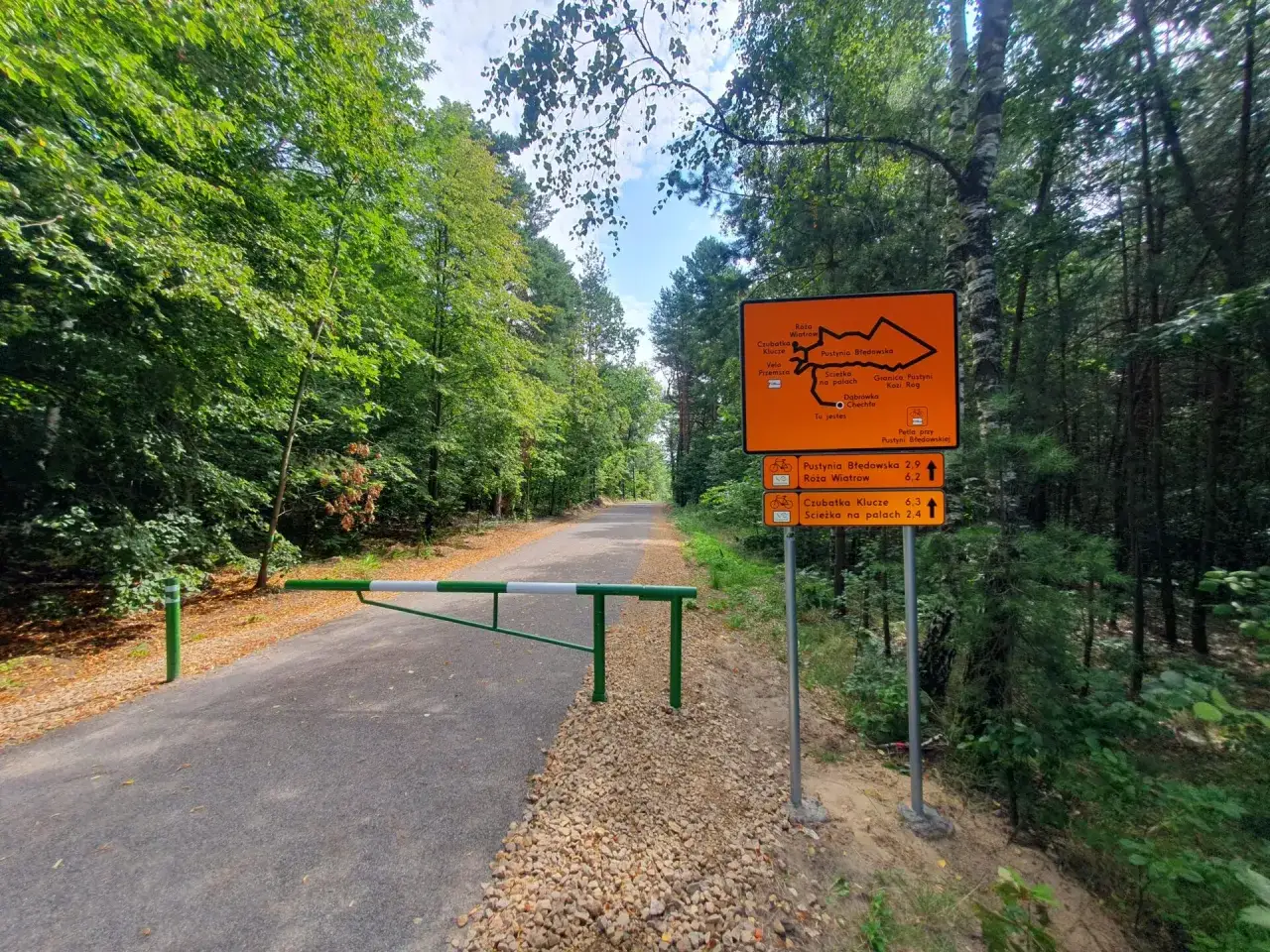 Pomarańczowy drogowskaz informuje o trasie rowerowej Pustynia Błędowska. Szlaban i leśna droga zapraszają do aktywnego wypoczynku.