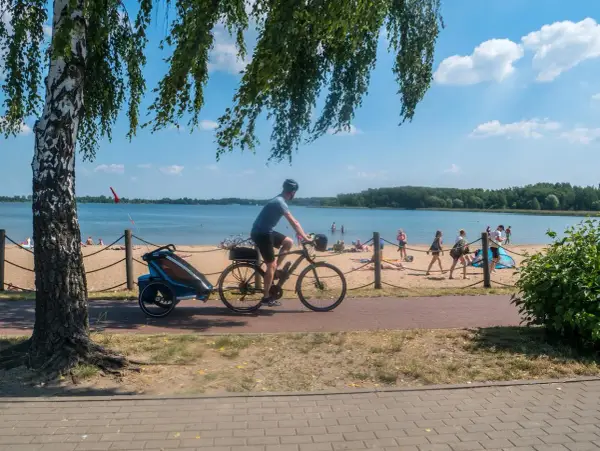 Rowerzysta z przyczepką jedzie ścieżką przy plaży nad Pogorią. Ludzie odpoczywają na piasku.